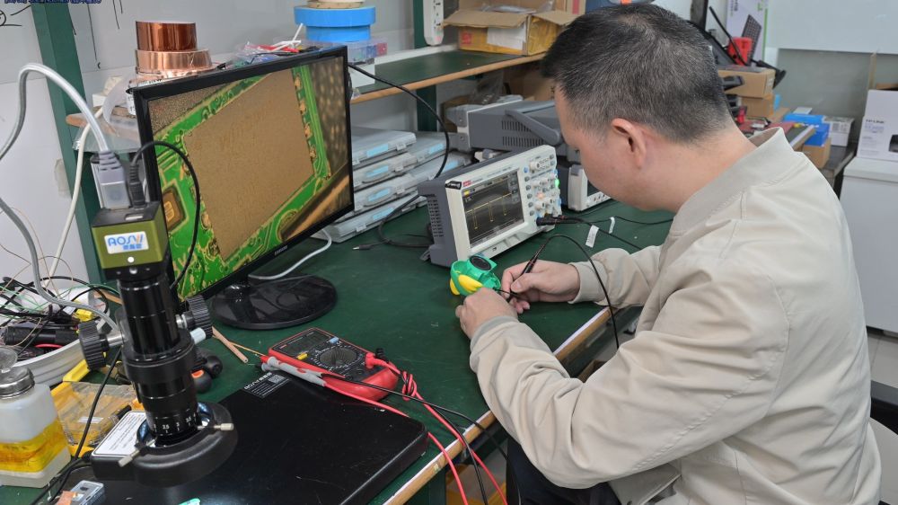 Engineer working on a voice-capture device at a bench with a digital microscope, oscilloscope, and multimeter